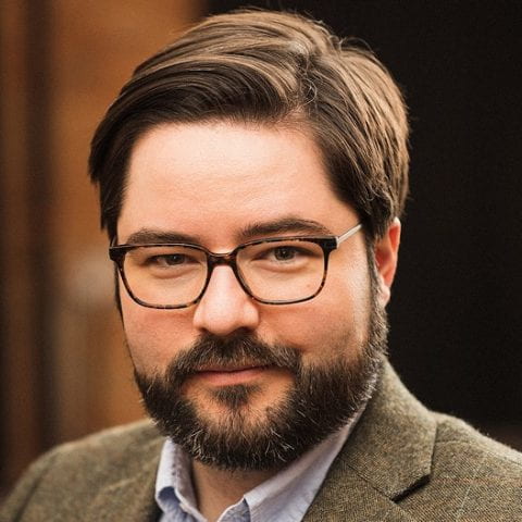 Trystan Goetze: a fair-skinned, male-presenting person with short dark hair and beard, wearing glasses and a tweed suit jacket.